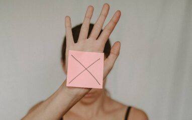 Frau hält rosa Post-it, auf dem ein Kreuz (Nein) zu sehen ist