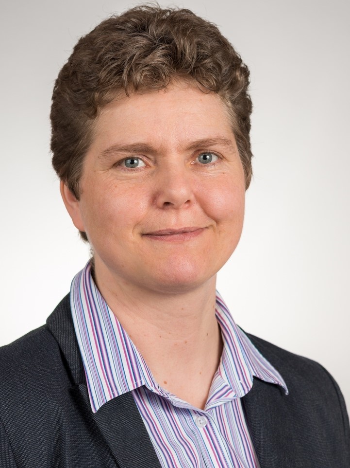 Portrait einer in die Kamera lächelnden Person. Sie trägt einen Blazer und eine gestreifte Bluse. Sie hat kurze Haare.